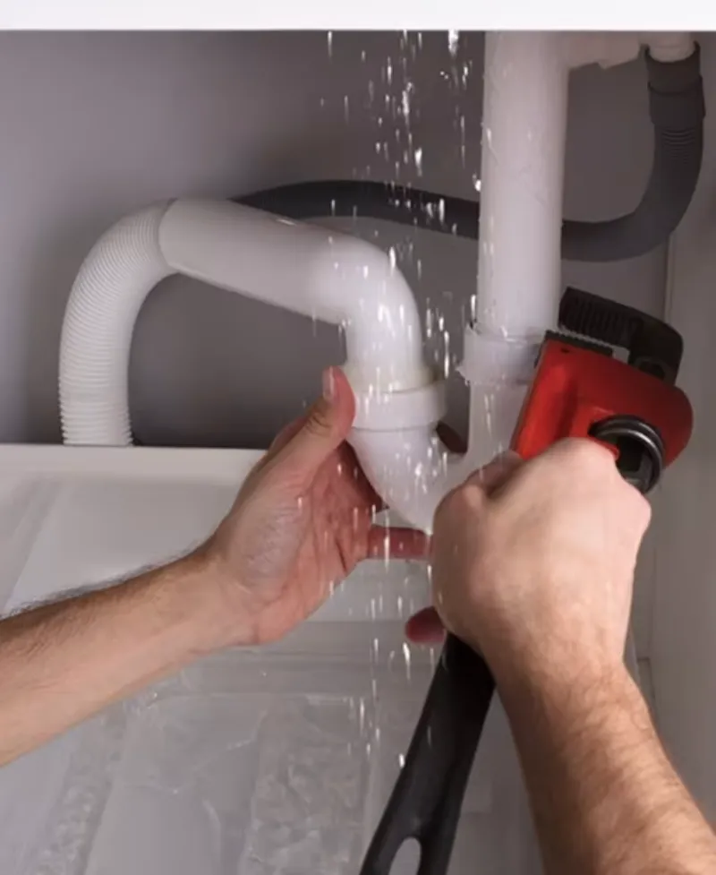 Hands tightening a leaking pipe joint under a sink with a pipe wrench